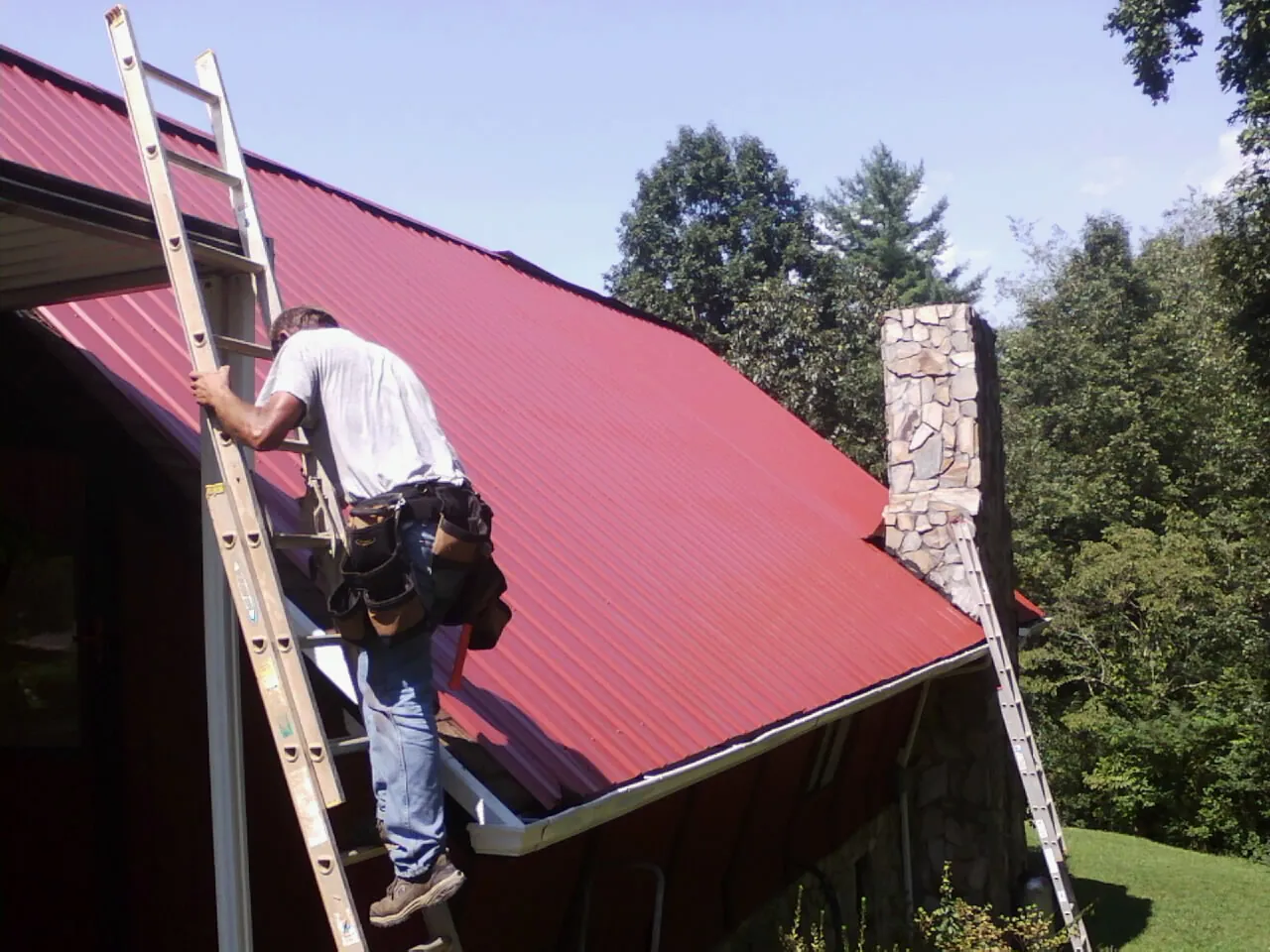 Roofer on a ladder beside a red metal roof and stone chimney — Soffit & Fascia Repair by Roofing Brothers in Newton Upper Falls