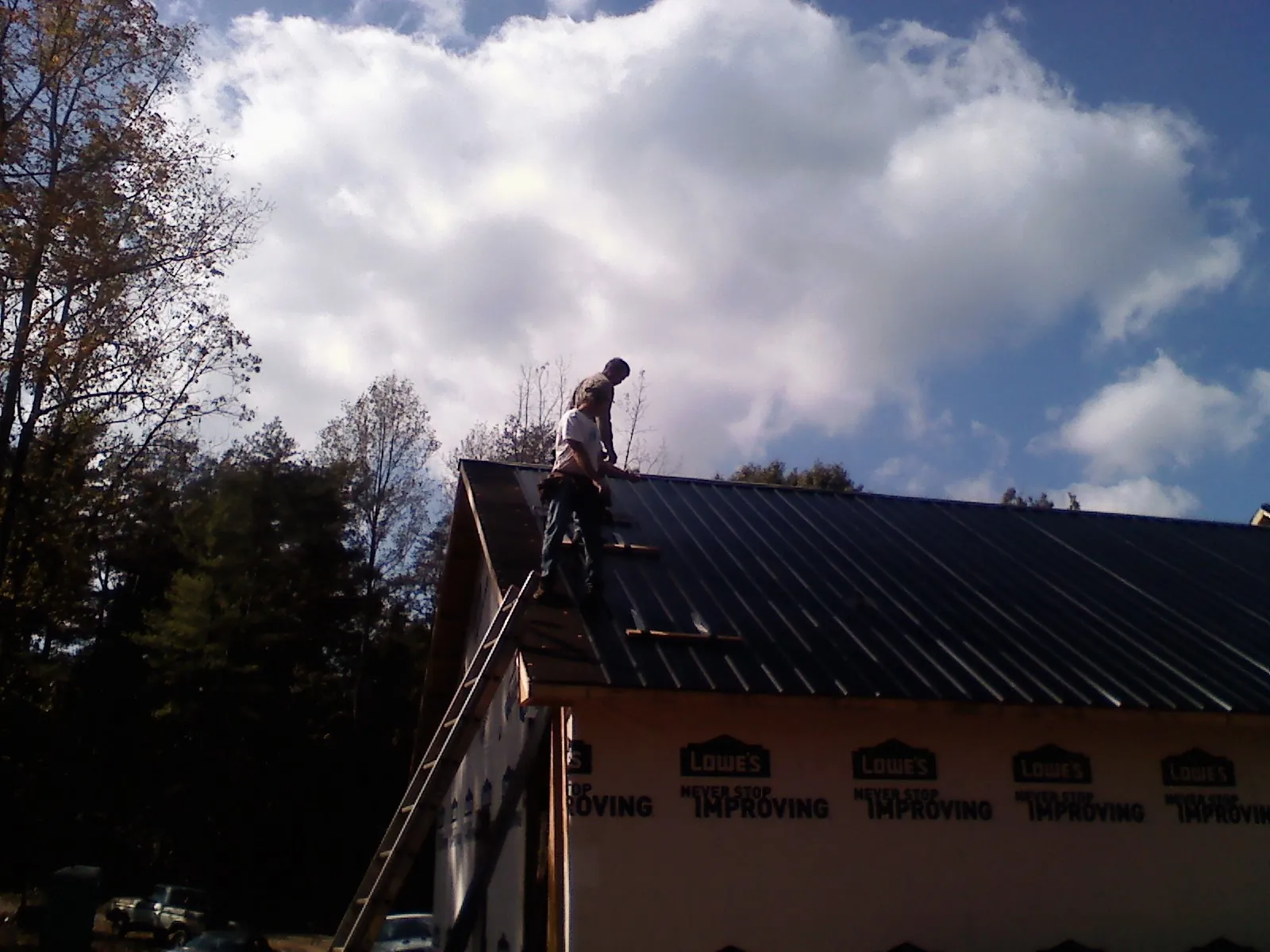 Roofer fastening dark metal panels high against a cloudy sky, from a Roof Inspection job in Newton Upper Falls